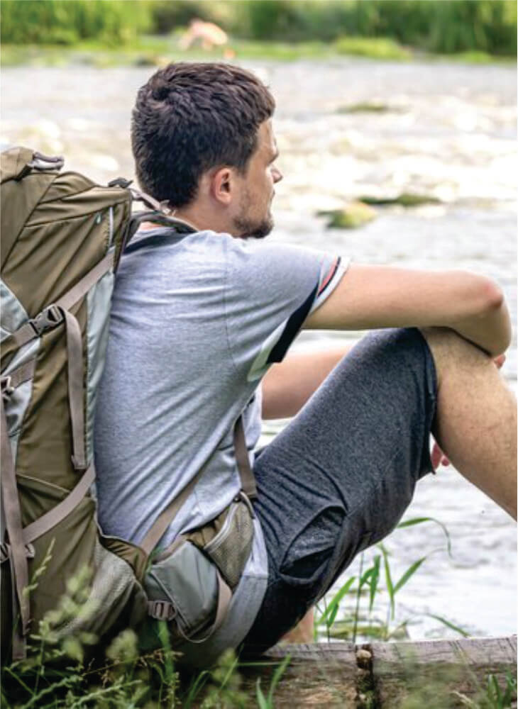 male budget traveler with a large backpack sits near Mara river on affordable 10 days Masai Mara camping safaris in Kenya