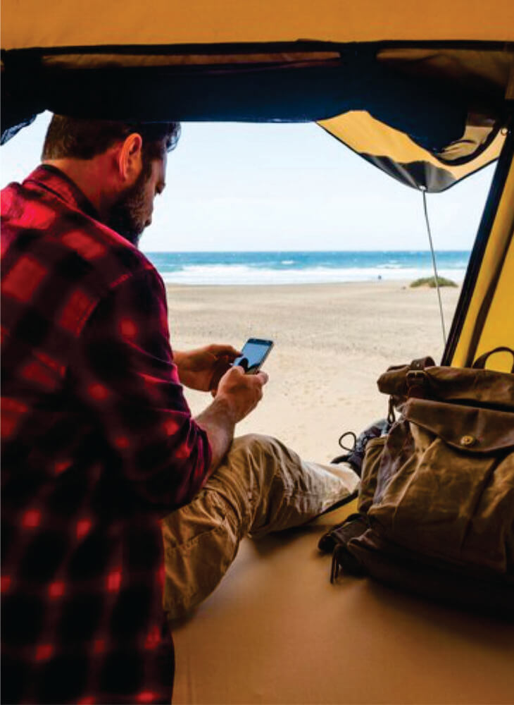 man sitting down inside a yellow tent near brown backpacking backpack and holding a phone on Kenya budget camping safari in Amboseli