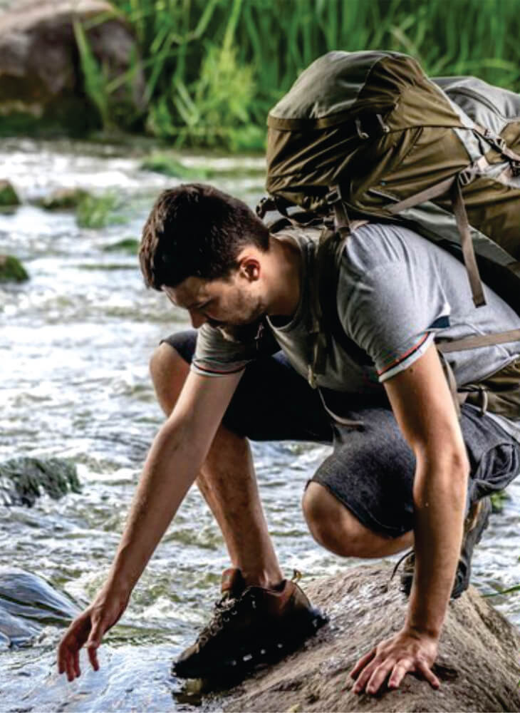 man with a large backpack cleaning his boots on the banks of Mara River on affordable Masai Mara camping safari in Kenya