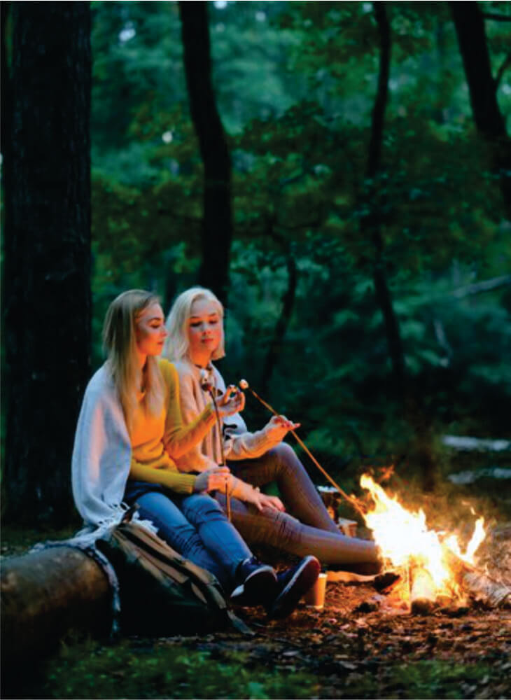 two ladies sit by bright bonfire at dusk near trees, spending nice time outdoors at a camping place on joining safaris in Masai Mara